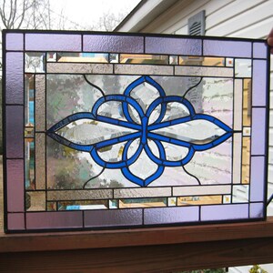 May include: Rectangular stained glass panel with a central blue and white floral design. The panel features a border of lavender and clear glass, with accents of gold and clear glass. The design is framed in black.