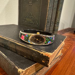 May include: A black and gold bangle watch with a floral design. The watch has a black face with gold hands and a gold bezel. The band is black with pink, blue, and white flowers. The watch is resting on a stack of old books.