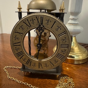 May include: Antique brass-colored clock with a round face featuring Roman numerals and fleur-de-lis markings. The clock's inner workings are visible, and it has a gold chain. The clock is set on a dark surface.
