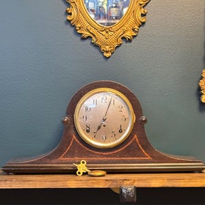 May include: An antique wooden mantel clock with a round face and gold-colored accents. The clock has a dark brown finish with decorative inlays and a brass-colored bezel. A gold-colored key and a small brass disc are on the wooden shelf in front of the clock.