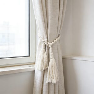 May include: Cream-colored curtains are held back by a white rope tieback with two large tassels. The curtains are made of a textured fabric and hang in front of a window. The tieback is knotted and adds a decorative element to the window treatment.