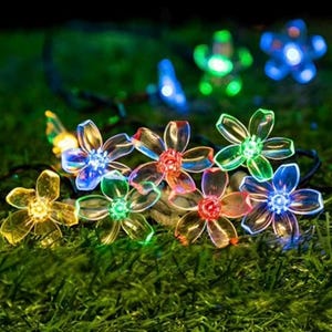 May include: A string of flower-shaped LED lights in various colors, including yellow, green, red, and blue. The lights are arranged on a black wire and are set against a backdrop of green grass, creating a decorative outdoor lighting display.