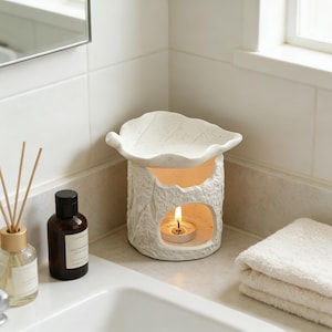 May include: A white ceramic essential oil diffuser with a leaf-shaped top and a textured, tree-bark-like body. A lit tea light candle glows inside. The diffuser sits on a bathroom counter next to a bottle and a folded white towel.