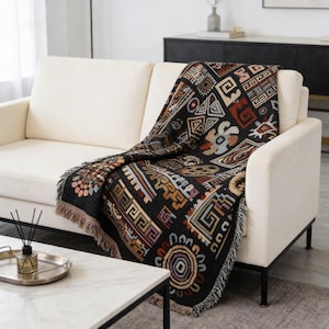 May include: A black throw blanket with a geometric pattern in shades of brown, beige, and rust is draped over a cream-colored sofa. The blanket has a fringed edge and a variety of shapes and designs.