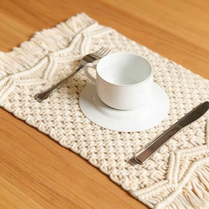 May include: A white teacup and saucer sit on a cream-colored macrame placemat, with a silver fork and knife. The placemat has a textured, woven design with fringe along the edges. The teacup has a gold rim.