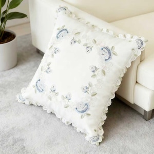 Puede incluir: Cojín decorativo blanco cuadrado con bordado floral azul y borde festoneado. El cojín está colocado sobre una alfombra gris claro junto a un sofá blanco y una planta en maceta.
