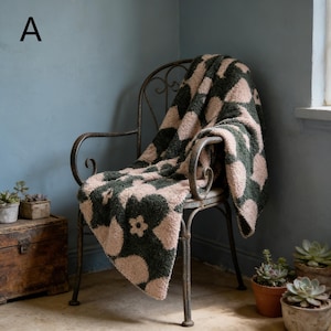 Puede incluir: Una manta suave con estampado floral, sobre una silla de metal vintage. El diseño floral es en verde oscuro y rosa claro. La silla está frente a una pared azul, con pequeñas plantas en macetas cerca.