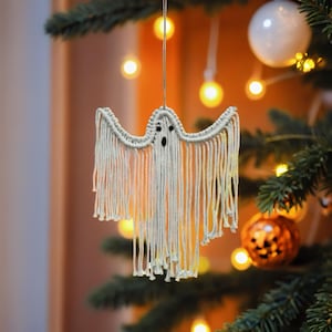 May include: A handmade, off-white macrame ghost decoration with a simple face, suspended from a string. The ghost hangs in front of a Christmas tree with warm, glowing lights and other decorations, creating a festive holiday scene.