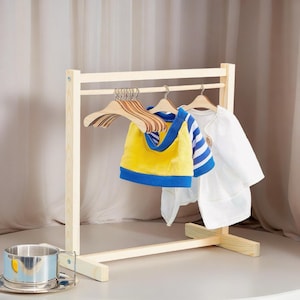 Może przedstawiać: Drewniany wieszak na ubrania z kilkoma ubraniami dla dzieci. Żółto-niebieska kamizelka, niebiesko-biała koszula w paski i biała koszula wiszą na drewnianych wieszakach. Srebrny kubek i spodek znajdują się na pierwszym planie.