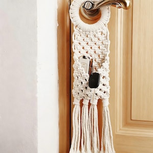 Könnte beinhalten: Ein cremefarbener Makramee-Tür-Organizer mit rundem Oberteil und rechteckiger Tasche. Die Tasche enthält ein Smartphone und Schlüssel. Der untere Teil des Organizers hat mehrere lange, herabhängende Quasten. Der Organizer hängt an einem goldfarbenen Türgriff.