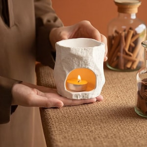 May include: A white ceramic candle holder with a textured surface. The holder has a cutout for a lit tealight candle, casting a warm glow. The candle holder is designed to hold a small, round candle. The overall theme is home decor.