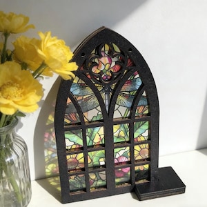 Puede incluir: Una ventana decorativa de madera negra con un diseño de vidriera que presenta una libélula y elementos florales. La ventana tiene una parte superior arqueada y una base rectangular. La vidriera tiene un diseño colorido y vibrante.