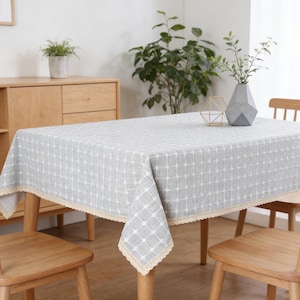 May include: A rectangular gray tablecloth with a white geometric pattern and a beige lace trim. The tablecloth is draped over a wooden table, with wooden chairs around it. A vase with greenery and a decorative object are on the table.