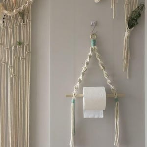 May include: A macrame toilet paper holder with a roll of white paper. The holder is made of off-white rope with a wooden dowel and teal accents. Other macrame wall hangings are also visible.