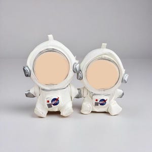 May include: Two white astronaut figurines with silver accents. Each figurine has a large, circular opening for a photo or image. The NASA logo is visible on the chest of each astronaut. The figurines are on a light gray background.
