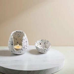 以下が含まれることがあります： 白い斑点模様のセラミックキャンドルホルダーと、中に火の灯ったキャンドル、小さなマッチングボウル。 キャンドルホルダーは卵のような形で、切り取られた部分があります。 ボウルとキャンドルホルダーには黒い斑点があります。