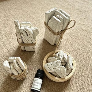 May include: Three bundles of porous, off-white volcanic stones tied with brown twine, alongside a small, round, light-colored stone bowl filled with loose volcanic stones and a small dark glass bottle with a white label and black cap.