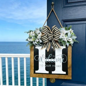 Könnte beinhalten: Ein hölzernes Willkommensschild mit einer schwarz-weiß gestreiften Schleife und floralen Akzenten. Das Schild zeigt das Wort "Welcome" in Schreibschrift und den Satz "TO OUR HOME" in fetter Schrift. Das Schild hängt an einer blauen Tür.