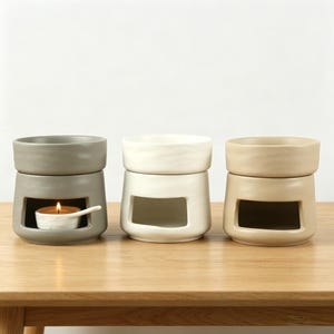 May include: Three ceramic oil burners in grey, white, and beige. Each burner features a cylindrical top bowl for wax or oil and a hollow base with a rectangular opening for a tea light candle. The grey burner contains a lit white tea light candle.