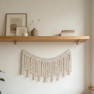 May include: A cream-colored macrame wall hanging with a geometric design and long fringe. The macrame is suspended from a wooden shelf, which also holds framed art, a vase with dried flowers, and books. The wall is painted a neutral color.