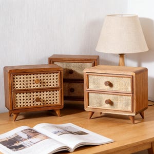 Può includere: Tre piccoli comodini in legno con frontali dei cassetti in rattan intrecciato e pomelli rotondi in legno. I comodini sono marroni e poggiano su gambe corte e angolate. Una lampada beige e una rivista aperta sono anche sulla superficie di legno.