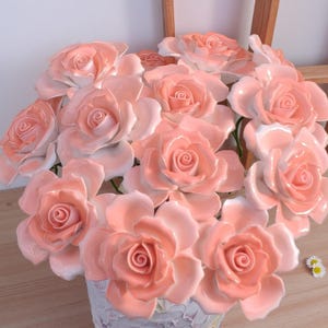 May include: A display of ceramic roses in pink and white hues. The roses feature intricate petal details and are presented in a decorative vase. The arrangement creates a soft, romantic floral display.