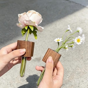May include: Two small, square wooden vases each hold a single flower. One vase holds a light pink peony, the other holds a cluster of white daisies with yellow centers. The vases have a glass tube for water.