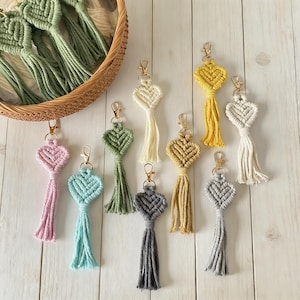 May include: A collection of handmade macrame heart-shaped keychains in various colors, including pink, blue, green, yellow, and gray. Each keychain features a gold-colored clasp and a long tassel. Some keychains are displayed in a woven basket.
