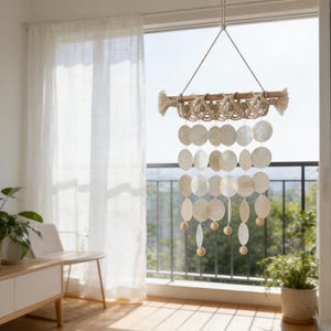 May include: A hanging wall decoration featuring a wooden rod with macrame detailing and a cascade of white, shell-shaped discs. The decor includes wooden beads and tassels, creating a natural, coastal aesthetic. The piece hangs in front of a window.