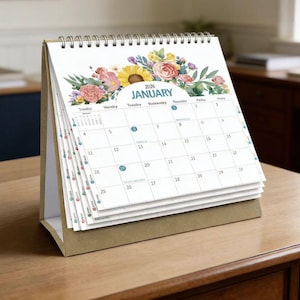 May include: A desk calendar for 2026, featuring a floral design with a sunflower and roses. The calendar is open to January, with the days of the week and dates visible. The calendar is on a wooden surface.