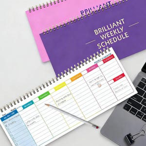 Könnte beinhalten: Ein Wochenplan-Notizbuch mit einem lila Einband mit dem goldenen Schriftzug "BRILLIANT WEEKLY SCHEDULE". Dahinter liegt ein rosa Notizbuch. Ein weißes Notizbuch mit Wochenplan ist mit einem Bleistift geöffnet.