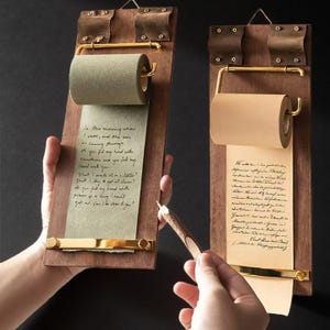 Könnte beinhalten: Zwei Holz-Notiztafeln mit Messingakzenten und Papierrollen. Eine Tafel hat grünes Papier, die andere braunes Papier. Die Tafeln haben eine Vintage-Ästhetik mit handschriftlichem Text auf dem Papier. Ein Bleistift wird in der Nähe einer der Tafeln gehalten.