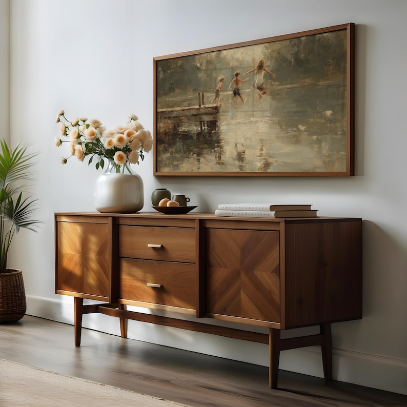 Puede incluir: Un aparador de madera de estilo moderno de mediados de siglo, con un cuadro enmarcado encima. El aparador tiene dos puertas y dos cajones con tiradores de lat&oacute;n. Un jarr&oacute;n con flores y objetos decorativos est&aacute;n encima. La pintura muestra a ni&ntilde;os saltando al agua.
