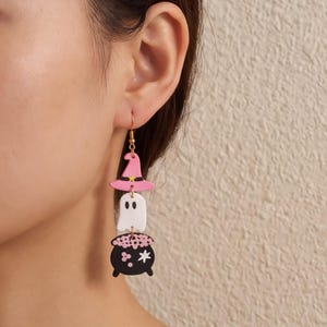 May include: A dangling earring featuring three stacked charms: a pink witch hat with a yellow star, a white ghost with black eyes, and a black cauldron filled with pink bubbling liquid and a white star, all suspended from a gold-toned hook.