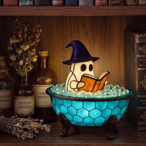 May include: A stained glass lamp shaped like a ghost wearing a dark purple witch hat and reading an open orange book. The ghost sits inside a turquoise blue, hexagonal-patterned bowl filled with clear glass beads, supported by three dark metal feet.