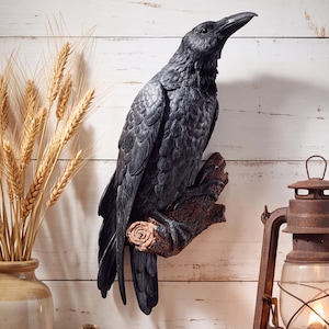 May include: A black raven figurine perched on a textured brown branch. The bird has detailed feather patterns and a sharp beak. The sculpture is mounted against a white wooden wall, positioned between a vase of dried wheat and a metal lantern.