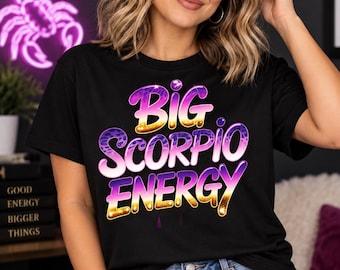 May include: A black short-sleeved t-shirt featuring the text "BIG SCORPIO ENERGY" in a stylized, gradient font with purple, pink, and gold colors. The lettering incorporates a subtle leopard print pattern and a glossy, reflective finish.