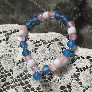 May include: A circular beaded bracelet featuring a repeating pattern of matte blue, translucent pink, and opaque white spherical beads. Small metallic silver spacer beads are interspersed between the larger colored beads along the elastic cord.