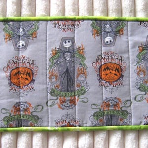 May include: A rectangular, quilted fabric item with a light gray background featuring a repeating pattern of Jack Skellington from The Nightmare Before Christmas. The design includes orange pumpkins and the text "Pumpkin King". The border is green.