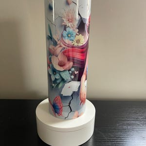 May include: A tall, insulated tumbler with a floral and butterfly design. The tumbler features a light blue background with cracks, and a design of pink, blue, and yellow flowers, and a butterfly. The tumbler is sitting on a white, round stand.