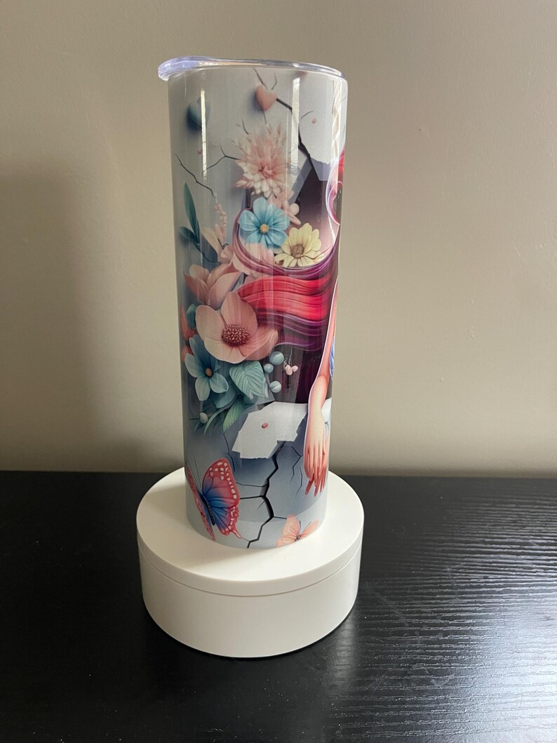 May include: A tall, insulated tumbler with a floral and butterfly design. The tumbler features a light blue background with cracks, and a design of pink, blue, and yellow flowers, and a butterfly. The tumbler is sitting on a white, round stand.