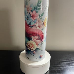 May include: A floral-themed tumbler with a white background, featuring pink, blue, and peach flowers, green leaves, and a yellow butterfly. The design includes flowing red hair and a cracked texture. The tumbler is cylindrical and has a clear lid.
