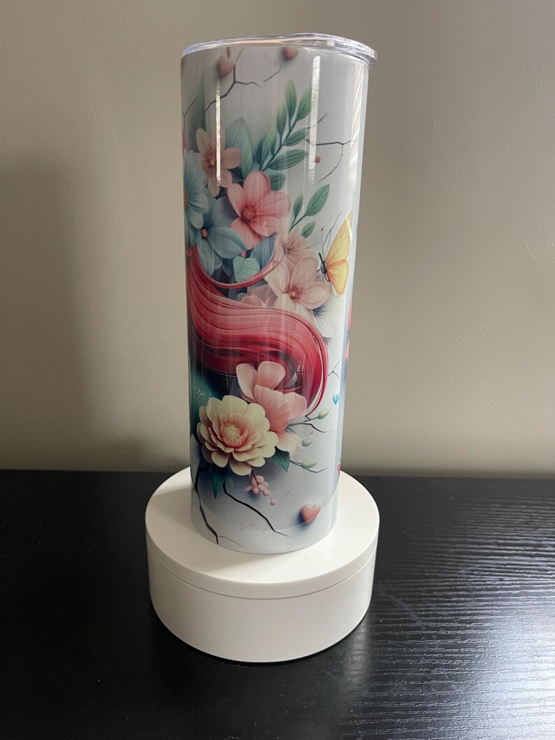 May include: A floral-themed tumbler with a white background, featuring pink, blue, and peach flowers, green leaves, and a yellow butterfly. The design includes flowing red hair and a cracked texture. The tumbler is cylindrical and has a clear lid.