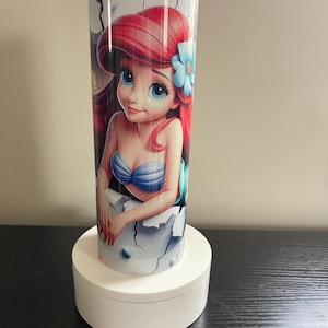 May include: A cylindrical tumbler featuring a vibrant illustration of a mermaid with red hair and a blue flower. The tumbler has a white background with a cracked design. The tumbler is approximately 8 inches tall.