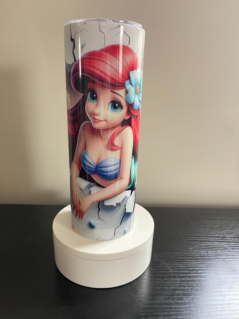 May include: A cylindrical tumbler featuring a vibrant illustration of a mermaid with red hair and a blue flower. The tumbler has a white background with a cracked design. The tumbler is approximately 8 inches tall.