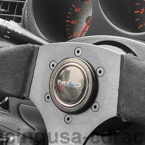 May include: A close-up of a black and grey steering wheel with a suede-like grip. The centre features a silver plate with the brand name "NRG". Dashboard gauges are visible in the background.