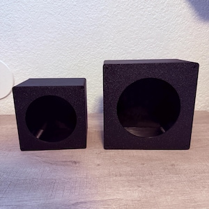 May include: Two black square speaker enclosures with circular cutouts on the front faces. The enclosures feature a textured matte finish and are positioned side by side, with the one on the right being larger than the one on the left.
