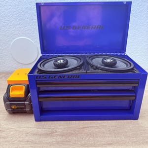 May include: A blue US GENERAL tool chest with two drawers and a top compartment, open to reveal two black speakers. The text "US GENERAL" is printed on the top and front of the chest. A yellow and black toy truck is to the left.
