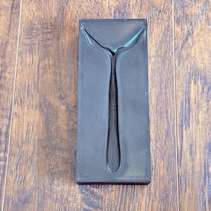 May include: A black, rectangular mold with a spoon-shaped cavity. The mold has a textured surface and is set against a wooden background. The spoon-shaped cavity is centered within the mold.