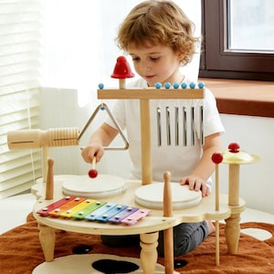 Puede incluir: Un juego de instrumentos musicales de juguete de madera con xilófono, tambores, platillos y un triángulo. El juego está pintado en varios colores y tiene una parte superior en forma de hongo. El niño está jugando con el juguete.